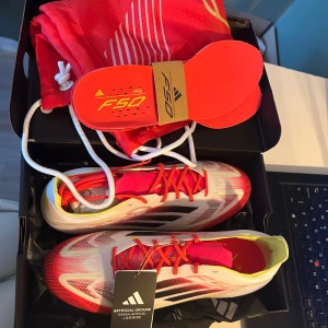 Adidas F50 Elite AG fotbollsskor 39 1/3 - Säljer ett par helt nya och oanvända Adidas F50 Elite AG fotbollsskor i vitt, svart, rött och gult. Skorna har snörning, platt sula med dobbar för konstgräs och en lätt, ventilerande ovandel i syntet. Snygg design med F50-detaljer och medföljande röd skopåse och extra innersulor. Kommer i orginalförpackning! Nypris 3099kr!
