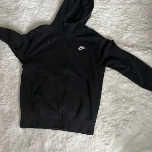 Svart Nike hoodie med dragkedja lite använd och smutsig  - Svart hoodie från Nike med vit logga på bröstet. Tröjan har huva, hel dragkedja och stora fickor framtill. Tillverkad i mjuk bomullsblandning, perfekt för chill eller streetwear. Klassisk och enkel design som funkar året runt.