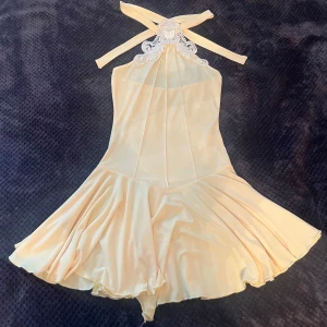 Champagnefärgad halterneckklänning S/M - Supersöt kort klänning i champagnefärgad satin med halterneck och broderad spetsdetalj vid halsen. Klänningen har en flowy kjol och knytband i nacken för en snygg passform. Perfekt för dig som vill sticka ut med en elegant och unik look.