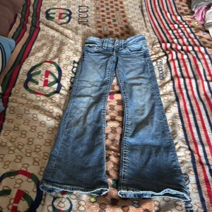 Blå bootcut jeans Gina Tricot 134 - Snygga blå jeans från Gina Tricot i storlek 134. Modellen har bootcut-ben och klassisk femficksdesign. Jeansen är i ett mjukt denimtyg med lätt tvättad look och normal passform. Perfekta för dig som gillar retrovibbar och bekväma jeans.