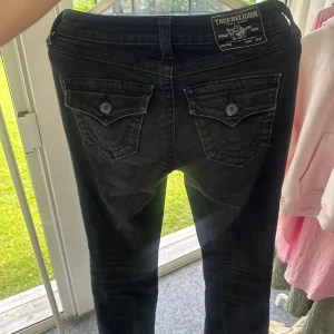 Svarta True Religion Becca Bootcut Jeans - Säljer ett par svarta True Religion Becca jeans med bootcut och midwaist. Snygga detaljer med markerade bakfickor och logga på bakre linningen. Jeansen är i stretchig bomull och har klassisk knappstängning framtill. Endast använda några få gånger och bara tvättade två gånger.