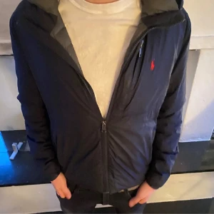 Mörkblå vindjacka från Polo Ralph Lauren - Snygg mörkblå vindjacka från Polo Ralph Lauren med huva och dragkedja framtill. Jackan har en röd broderad logga på bröstet och två fickor med dragkedja. Perfekt för kyliga dagar och enkel att matcha med det mesta.