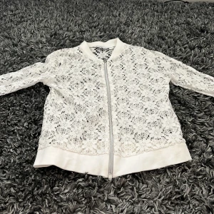 Vit spets bomberjacka från Mywear Young - Supersöt vit bomberjacka från Mywear Young med blommig spets över hela jackan. Jackan har dragkedja framtill, ribbade muddar och lång ärm. Perfekt för dig som vill ha en unik och fräsch look med extra detaljer.
