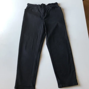 Svarta byxor från Lindex, stl 146 - Svarta byxor från Lindex i storlek 146 med resår i midjan och raka ben. Klassisk modell med fickor bak och bälteshällor. Perfekta till vardags och lätta att matcha med olika stilar.