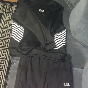 Svart EA7 långärmad tröja med ränder - Svart långärmad tröja från EA7 Emporio Armani med vita horisontella ränder på sidorna och logga på bröstet. Tröjan har en sportig vibe och är gjord i ett glansigt, mjukt material som känns skönt mot huden.