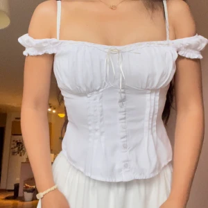 Vit korsett-topp med snörning - Superfin vit korsett-topp med romantiska puffärmar som kan bäras både off-shoulder och på axlarna. Toppen har knappar framtill, markerade sömmar och söt snörning vid bysten. Figurnära passform och lätt rynkade detaljer ger en feminin vibe.