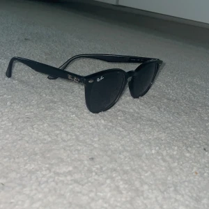 Svarta Ray-Ban Wayfarer solglasögon - Klassiska svarta Ray-Ban Wayfarer solglasögon med ikonisk fyrkantig form och silvriga detaljer vid bågarna. Snygg och tidlös design med Ray-Ban-logga på både lins och skalmar. Perfekta för dig som vill ha en cool och stilren look. Ganska slitna 