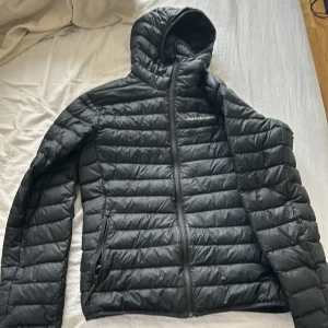 Svart dunjacka från Peak Performance - Svart dunjacka från Peak Performance med huva och dragkedja framtill. Jackan är quiltad med horisontella sömmar och har två fickor med dragkedja. Perfekt för kalla dagar och enkel att matcha med allt. Bara använt den några gånger men känner att de inte riktigt är jag 