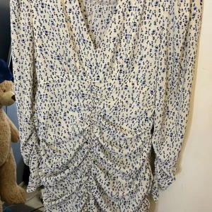 Blommig kortklänning med rynk - Supersöt kortklänning med små blå blommor på vit botten. Klänningen har långa ärmar, v-ringning och snygga rynkdetaljer framtill som ger en cool siluett. Perfekt för dig som gillar romantiska och trendiga vibes.