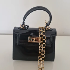 Svart minihandväska med guldkedja - Snygg svart minihandväska i glansigt material med guldfärgad kedja och vridlås framtill. Väskan har ett stilrent och modernt uttryck med ett stadigt handtag och detaljer i guld. Perfekt accessoar för att lyfta din outfit.