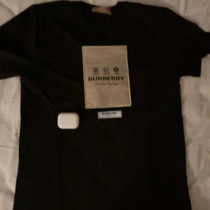 Svart Burberry t-shirt med beige patch - Svart t-shirt från Burberry med en stor beige patch på bröstet där Burberry-loggan och texten 'London England' syns. Klassisk passform och korta ärmar. Perfekt för dig som gillar stilrena designer och vill sticka ut med ett exklusivt märke.