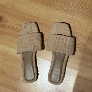 Beige sandaler oanvända  - Snygga beige sandaler med fyrkantig tå och bred rem i textil med strukturmönster. Platt sula och öppen design, perfekta för varma dagar. Stilren look som passar till många outfits. Endast använd 1 gång inomhus. 