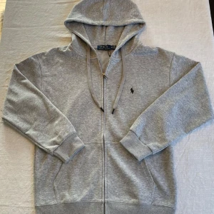 Grå hoodie från Polo Ralph Lauren - Säljer en klassisk grå hoodie från Polo Ralph Lauren med dragkedja och huva. Tröjan har ribbade muddar, två fickor fram och den ikoniska broderade loggan på bröstet. Perfekt för en avslappnad stil.