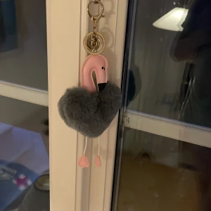 Grå fluffig flamingo-nyckelring från NYATU - Söt nyckelring i form av en flamingo med rosa detaljer och fluffig grå kropp. Perfekt som accessoar till väskan eller nycklarna. Flamingon har små ben med rosa fötter och ett guldigt nyckelringsfäste.