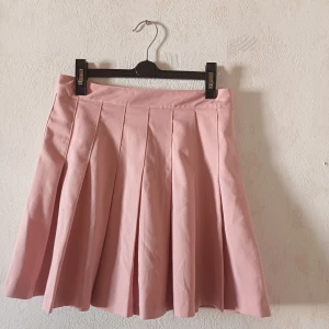 Rosa plisserad kjol - Säljer en söt rosa plisserad kjol med knappdetalj i midjan. Kjolen har en kort längd och är perfekt för en trendig look. Den är gjord i ett mjukt material som ger en fin passform.