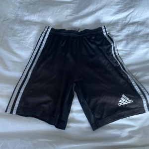 Svarta träningsshorts från Adidas - Säljer ett par svarta träningsshorts från Adidas med de klassiska tre vita ränderna längs sidorna. Shortsen har en elastisk midja och är perfekta för sport och träning. Adidas-loggan är tryckt på ena benet.