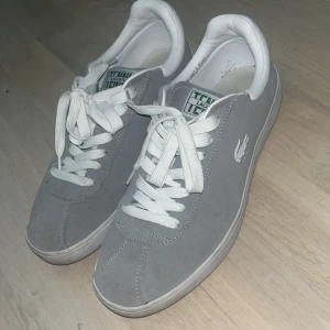 Grå sneakers från Lacoste - Snygga grå sneakers från Lacoste med vit snörning och rund tå. Skorna har en klassisk design med Lacoste-loggan på sidan och är perfekta för en avslappnad stil.