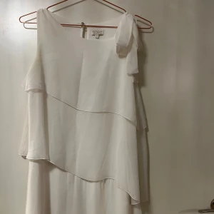 Vit lång klänning med volang - Säljer en vit, axelbandslös klänning från Mimirose med asymmetrisk design och volangdetaljer. Klänningen har en oneshoulder-modell med en dekorativ rosett på axeln och är tillverkad i ett lätt, flödande material.