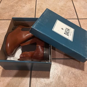 Bruna chelsea boots från Dasia - Säljer ett par snygga bruna chelsea boots från Dasia med croco-detaljer på hälen och resår på sidorna. Skorna har rund tå och låg klack, perfekt för dig som vill ha en stilren look med lite edge.  Pris kan diskuteras vid smidig affär:)