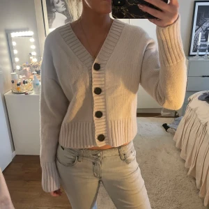 Beige stickad kofta med stora knappar - Mysig beige stickad kofta i kashmir och ull med stora svarta knappar framtill och bred ribbad kant. Koftan har lång ärm och croppad passform, perfekt att slänga på sig över ett linne eller t-shirt. 