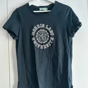 Mörkblå t-shirt från Morris Lady med vit tryckt text och emblem på bröstet. Klassisk rund halsringning och korta ärmar. Perfekt för en avslappnad och stilren look.