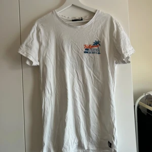 Vit t-shirt med tryck från SALIGIA - Vit t-shirt från SALIGIA med färgglatt tryck 'Hollywood Burger Station' och palm på bröstet och ryggen. Klassisk rund hals och korta ärmar. Tillverkad i 100% bomull för en skön känsla.