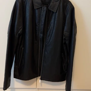 Svart skinnjacka med dragkedja - Säljer en stilren svart skinnjacka med dragkedja framtill och klassisk krage. Jackan har långa ärmar och två fickor med dragkedja på sidorna. Perfekt för dig som vill ha en tidlös look.
