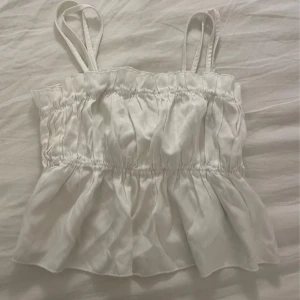 Vit ärmlös topp med volangdetaljer - Säljer en söt vit ärmlös topp med dubbla axelband och volangdetaljer. Toppen har en luftig och lätt design, perfekt för varma dagar. Aldrig använd!!  