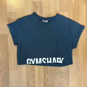 Svart croppad t-shirt från Gymshark - Säljer en svart croppad t-shirt från Gymshark med vit logga längst ner. T-shirten har korta ärmar och en avslappnad passform, perfekt för träning eller gymmet.