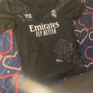 Real Madrid svart fotbollströja Y-3 Bellingham - Svart Real Madrid fotbollströja med Y-3 logga och Emirates Fly Better-tryck på framsidan. Snygga rosdetaljer i mönstret och Real Madrid-emblem på bröstet. Baksidan har Bellingham och nummer 5 med en stor vit siluett.