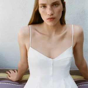 Vit topp med smala axelband - Säljer en så söt och stilren vit topp från Zara med smala axelband. Toppen har en markerad midja och knappar framtill. Perfekt för varma dagar och enkel att matcha! Storlek M/38 och prislappen sitter kvar.