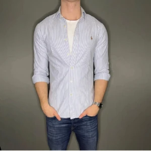 Vit och blå randig skjorta från Ralph Lauren - Säljer en stilren vit och blå randig skjorta från Ralph Lauren. Skjortan har långa ärmar och knappar framtill. Perfekt för en klassisk och avslappnad look. 
