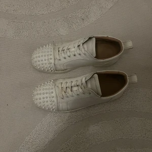 Vita sneakers med nitar från Christian Louboutin - Snygga vita sneakers från Christian Louboutin med coola nitar på tån. Skorna har snörning och en klassisk rund tå. Perfekta för att ge din outfit en edgy touch.