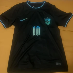 Svart Brasilien fotbollströja Neymar Jr 10 - Säljer en svart Brasilien fotbollströja från Nike med ljusblå detaljer. Tröjan har Neymar Jr och nummer 10 på ryggen, samt Brasilien-emblem och Nike-logga på bröstet. Tillverkad i Dri-Fit material för bra komfort.