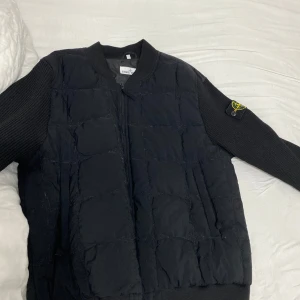 Svart quiltad jacka från Stone Island - Säljer en svart quiltad jacka från Stone Island med ribbade ärmar och dragkedja framtill. Jackan har den klassiska loggan på vänster ärm och en stilren, sportig look. Perfekt för dig som gillar streetwear och vill ha något snyggt och bekvämt.