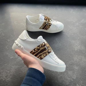 Valentino Rockstud  - Säljer ett par vita Valentino Rockstud sneakers med leopardmönstrad detalj och guldiga nitar på sidorna. Skorna har platt sula och rund tå, samt klassisk snörning. Perfekta för dig som vill sticka ut med både lyx och edge.Box och Dustbag inngår