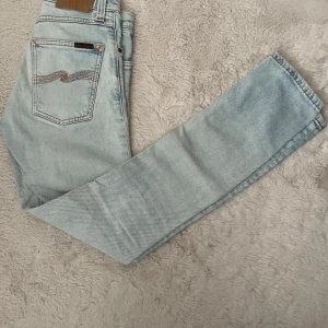 Ljusblå jeans från Nudie Jeans - Säljer ett par ljusblå jeans från Nudie Jeans med klassisk femficksdesign och orangea sömmar.  Riktigt feta nu inför sommaren! Jeansen har raka ben och normal passform, perfekt för en avslappnad stil. Märkeslogga på bakfickan.