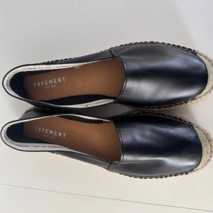Svarta espadrillos från Pavement - Snygga svarta espadrillos från Pavement med ovandel i skinn och klassisk flätad sula i naturfärgat material. Skorna har rund tå och platt sula, perfekta för en avslappnad och stilren look. Helt oanvända nypris 1100kr