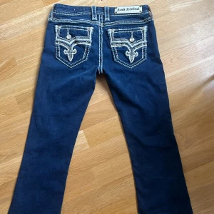 Jätte fina Rock Revival jeans med broderade bakfickor - Jätte snygga mörkblå bootcut lowwaist jeans från Rock Revival med vita kontrastsömmar och detaljerade broderier. Säljer för att dom är för korta❤️ Mått: Midja 36, Innerben 68, längd 100, benöppning 22. Priset går att diskutera!!