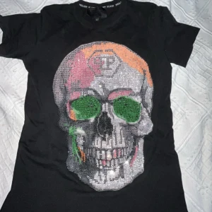 Svart t-shirt med dödskalle från Philipp Plein - Snygg svart t-shirt från Philipp Plein med en stor, färgglad dödskalle i paljetter på framsidan. Perfekt för dig som vill sticka ut med en unik stil. T-shirten har en normal passform och korta ärmar.