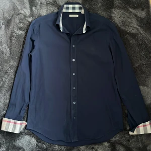 Burberry skjorta - Säljer en marinblå skjorta från Burberry med klassiskt rutigt mönster på insidan av kragen och manschetterna. Mycket bra skick utan defekter eller liknande. Ny pris runt 4000kr mitt pris 900kr, pris kan diskuteras vid snabb affär. Mvh Viktor🙌