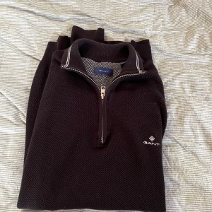 Svart stickad halvzip-tröja från GANT - Säljer en svart stickad tröja från GANT med halv dragkedja och vit broderad logga på bröstet. Tröjan har hög krage med vit detaljsöm och är perfekt för lager-på-lager-stil. Klassisk och stilren modell. Den har två defekter men är lätt lagade. Priset går därför att diskuteras👍😁