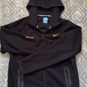 Nike Nocta Tech Fleece - Svart Tech Fleece Nike NOCTA med gul broderad logga och text på bröstet. Jackan har dragkedja framtill, huva och fickor med dragkedja. Snygga detaljer i gult även på ryggen. Perfekt för en sportig och stilren look.