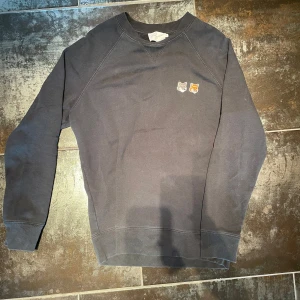 Maison Kitsuné sweatshirt - Säljer en stilren grå sweatshirt från Maison Kitsuné med två broderade rävhuvuden på bröstet. Tröjan har rund halsringning och långa ärmar. Perfekt för dig som gillar minimalistisk design med en twist.