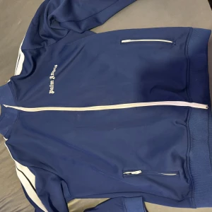 Blå träningsjacka från Palm Angels - Snygg blå träningsjacka från Palm Angels med vita detaljer och dragkedja. Jackan har en broderad logga på bröstet och ribbade muddar. Perfekt för en sportig look.