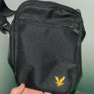 Lyle & skott väska - Snygg och praktisk svart axelväska med en gul logotyp framtill. Väskan har flera fack med dragkedja för enkel organisering och en justerbar axelrem för bekväm användning. Perfekt för att bära med sig det viktigaste på språng.