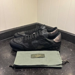 Svarta Valentino Rockrunner sneakers - Snygga svarta Valentino Rockrunner sneakers med ett diskret camouflage-mönster. Skorna har en kombination av skinn och mocka med snörning och en rund tå. Perfekta för en stilren look.