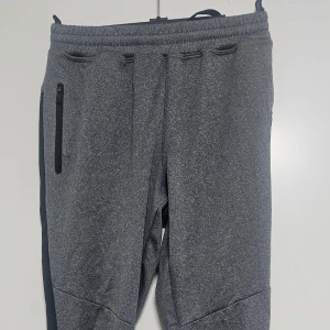 Grå mjukisbyxor från SOC - Grå mjukisbyxor från SOC med svarta detaljer längs sidorna och dragkedjeficka på ena benet. Byxorna har resår i midjan och är tillverkade i ett mjukt syntetmaterial som andas. Perfekta för träning eller chill.