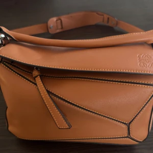 A+ Brun axelväska från Loewe - Snygg brun axelväska från Loewe i skinn med unika geometriska sömmar och en praktisk dragkedja. Väskan har en justerbar axelrem och en stilren logotyp präglad på framsidan. Perfekt för att ge din outfit en lyxig touch.