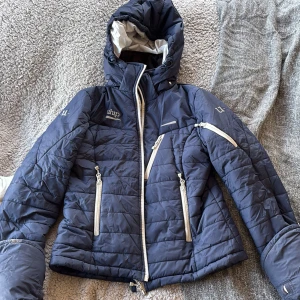 Blå jacka från Uhip - Säljer en snygg blå jacka från Uhip med dragkedjor och huva. Jackan har flera praktiska fickor med dragkedja och är perfekt för kyligare dagar. Den är bekväm och varm. Modell Ice och storlek 36. Endast använd en vinter, nypris 2295kr